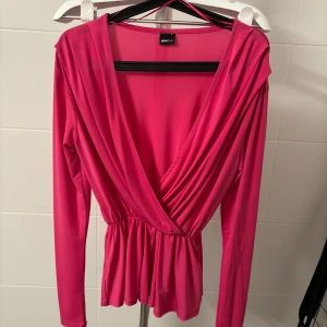 Rosa omlottopp från Gina Tricot - Snygg rosa omlottopp från Gina Tricot med långa ärmar och v-ringning. Toppen har en elastisk midja som ger en smickrande passform. Perfekt för en chic och bekväm look.