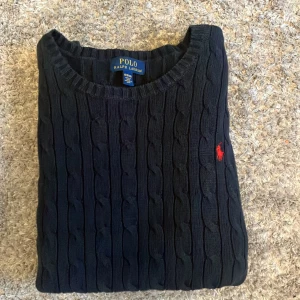 Marinblå kabelstickad tröja från Polo Ralph Lauren - Säljer en stilren marinblå kabelstickad tröja från Polo Ralph Lauren med det klassiska röda logotypbroderiet på bröstet. Tröjan har långa ärmar och en rund halsringning. Perfekt för en elegant och avslappnad look. Storlek XL i barn så var storlek S-M