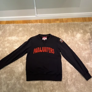 Svart fleece tröja från Parajumpers - Säljer en svart fleece tröja från Parajumpers med röd text på bröstet och ett märke på ärmen. Tröjan har långa ärmar och en rund halsringning. Perfekt för kyliga dagar.