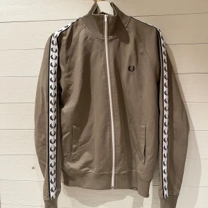 Beige track jacket med logotypband - Snygg olivgrön track jacket från Fred Perry med vita logotypband längs ärmarna. Jackan har en dragkedja framtill och ribbade muddar vid ärmslut och nederkant. Storlek M. Skriv för fler frågor. :)