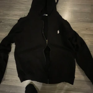 Ralph Lauren Zip Hoodie  - Snygg svart huvtröja från Ralph Lauren med dragkedja och broderad logga på bröstet. Perfekt för en avslappnad stil. Huvan ger extra komfort och värme.