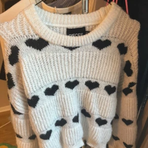 Vit stickad tröja med hjärtmönster från Pieces - Mysig vit stickad tröja från Pieces med svarta hjärtan. Perfekt för en avslappnad stil med en söt touch. Tröjan har en rund hals och långa ärmar.