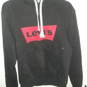 Svart hoodie från Levi's - Säljer en svart hoodie från Levi's med röd logga på bröstet. Den har en justerbar huva med gråa snören och en stor ficka framtill. Perfekt för en avslappnad stil.