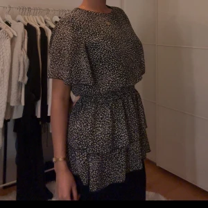 Svart och vit prickig blus - Snygg svart och vit prickig blus med korta ärmar och volangdetaljer. Klänningen har en elastisk midja som ger en fin passform och en luftig känsla. Perfekt för en stilren look.