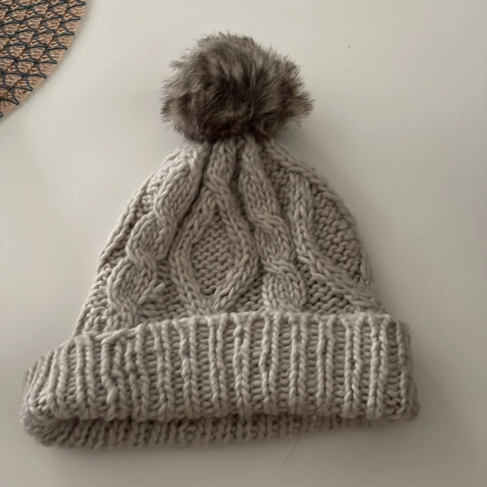 Säljer en mysig grå stickad mössa med en fluffig pälsboll på toppen. Mössan har ett klassiskt flätmönster och en bred ribbad kant som ger extra värme. Perfekt för kalla vinterdagar!. Asusteet.