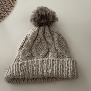 Grå stickad mössa med pälsboll - Säljer en mysig grå stickad mössa med en fluffig pälsboll på toppen. Mössan har ett klassiskt flätmönster och en bred ribbad kant som ger extra värme. Perfekt för kalla vinterdagar!