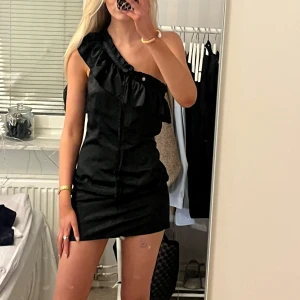 Svart oneshoulder klänning - Snygg svart oneshoulder klänning med volangdetaljer. Perfekt för en kväll ute eller en speciell tillställning. Den korta längden ger en chic look. Går att knyta rosett där fram eller stoppa in snörena.