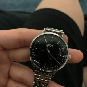 Svart klocka från Regal - Stilren klocka från Regal med svart urtavla och silverfärgad länkarmband. Klockan har en minimalistisk design med tydliga visare och indexmarkeringar. Perfekt för en elegant och tidlös look.