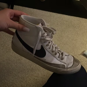 Nike Blazer i vitt och svart - Säljer ett par klassiska Nike Blazer sneakers i vitt med svart swoosh. Skorna har hög modell med snörning och är tillverkade i skinn. Perfekta för en stilren look. Kan sänka priset vid snabb affär