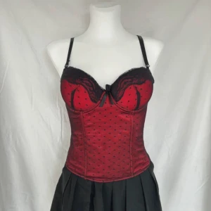 Röd korsett med spetsdetaljer från Feeling - Snygg röd korsett från Feeling med svarta spetsdetaljer och prickigt mönster. Den har justerbara axelband och hakknäppning bak för perfekt passform. Perfekt för en elegant look.