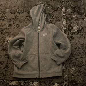 Säljer en klassisk grå hoodie från Nike med dragkedja och huva. Tröjan har långa ärmar och en liten Nike-logga på bröstet. Perfekt för en avslappnad stil. 