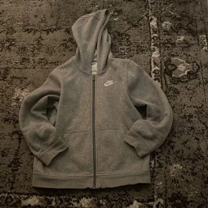 Grå hoodie från Nike storlek 150 - Säljer en klassisk grå hoodie från Nike med dragkedja och huva. Tröjan har långa ärmar och en liten Nike-logga på bröstet. Perfekt för en avslappnad stil. 