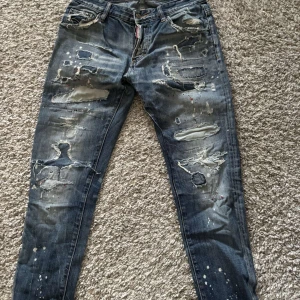 DSQUARED2 blå - Säljer ett par blå jeans med en cool, sliten look och flera revor. De har en klassisk femficksdesign och en knappgylf. Perfekta för en avslappnad stil.