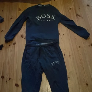 Hugo Boss dress - Säljer ett set med mörkblå tröja och matchande byxor från Hugo Boss. Tröjan har långa ärmar och ett stort BOSS-tryck på framsidan. Byxorna har en elastisk midja med snörning och ett mindre BOSS-tryck på benet. Perfekt för en avslappnad stil. Setet är i strl M