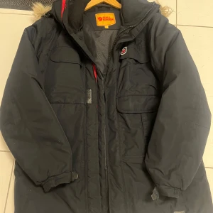 Svart parkajacka från Fjällräven - Säljer en svart parkajacka från Fjällräven med pälsdetaljer på huvan. Jackan har dragkedja och flera praktiska fickor framtill. Perfekt för kalla dagar.