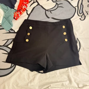 Svarta shorts med knappar från Gina Tricot - Snygga svarta shorts från Gina Tricot med dekorativa guldknappar framtill. De har en dragkedja på sidan för enkel på- och avtagning. I storlek 38 men liten i storleken. 