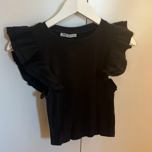 Svart topp med volangärmar från Zara - Snygg svart topp från Zara med volangdetaljer på ärmarna. Perfekt för att ge en extra touch till din outfit. Toppen har en rund halsringning och är tillverkad i ett mjukt material.