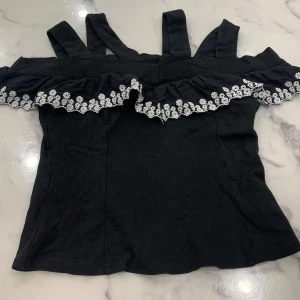 Svart topp med volang och blommig spets från Lost Ink - Snygg svart topp från Lost Ink med volangdetalj och blommig spets längs kanten. Perfekt för en stilren look med en touch av romantik. Axelbandslös design som ger en elegant känsla.