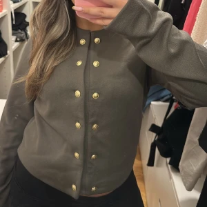 Grön kofta från H&M - Säljer en stilren grå kofta från H&M med guldknappar framtill. Koftan har långa ärmar och en rund halsringning, perfekt för en elegant look. Passar bra till både vardag och mer uppklädda tillfällen. Grön, mörkgrön, armégrön 