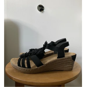 Sandaler med blomdetalj - Säljer dessa supersnygga sandaler med kilklack från Rieker. De är gjorda i äkta läder och har väldigt fina blomdetaljer. Stl 36. Fint skick, använda fåtal gånger. Perfekt för semestern. Skriv om du har frågor✨