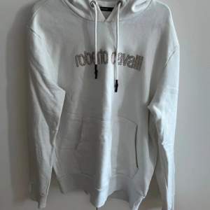 Säljer en snygg vit hoodie från Roberto Cavalli. Den är i storlek M och har en stor ficka på magen. Hoodien har långa ärmar och en justerbar huva med dragsko. Perfekt för en avslappnad och stilren look!