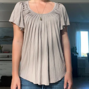 Beige blus med korta ärmar - Säljer en fin beige blus med korta ärmar. Den har en lös passform och är gjord av ett mjukt och bekvämt material, vilket gör den skön att ha på sig hela dagen. Den har också fina veckdetaljer vid halsringningen som ger den en extra touch av stil. Passar perfekt till jeans eller kjol!