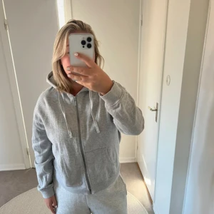 Grå hoodie med dragkedja - Säljer en mysig grå hoodie med dragkedja framtill. Den har en luva med snören och långa ärmar. Perfekt för en avslappnad look eller för att hålla sig varm under kyliga dagar. Passar bra till både vardag och träning.
