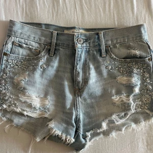 Ljusa jeansshorts från Abercrombie & Fitch - Säljer ett par snygga lågmidjade jeansshorts från Abercrombie & Fitch i storlek W26/S. De har en cool distressed look med slitningar och fransar längs kanterna. Shortsen är dekorerade med broderade blommor och små pärlor på framsidan, köpta här på plick men tyvärr lite för små i midjan så säljer därför vidare! 🥰💕💕 kan gå ner i pris vid snabb affär🥰