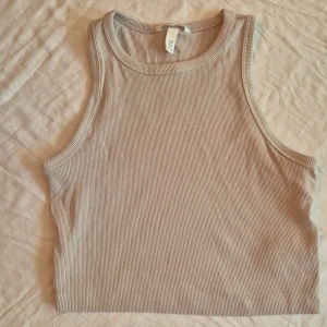 Beige ribbad linnetopp från H&M - Säljer en snygg beige ribbad linnetopp från H&M. Toppen är ärmlös och croppad, perfekt för sommaren eller som en del av en lager-på-lager outfit. Den är i nyskick och har en tight passform som framhäver figuren. Superbekväm och stilren!