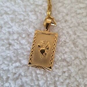 Guldhalsband med rektangulär hängsmycke - Säljer ett elegant guldhalsband med en rektangulär hängsmycke. Hängsmycket har ett detaljerat mönster med en ros i mitten och en vacker kantdesign. Perfekt för att ge en touch av lyx till din outfit. Passar både till vardags och fest.