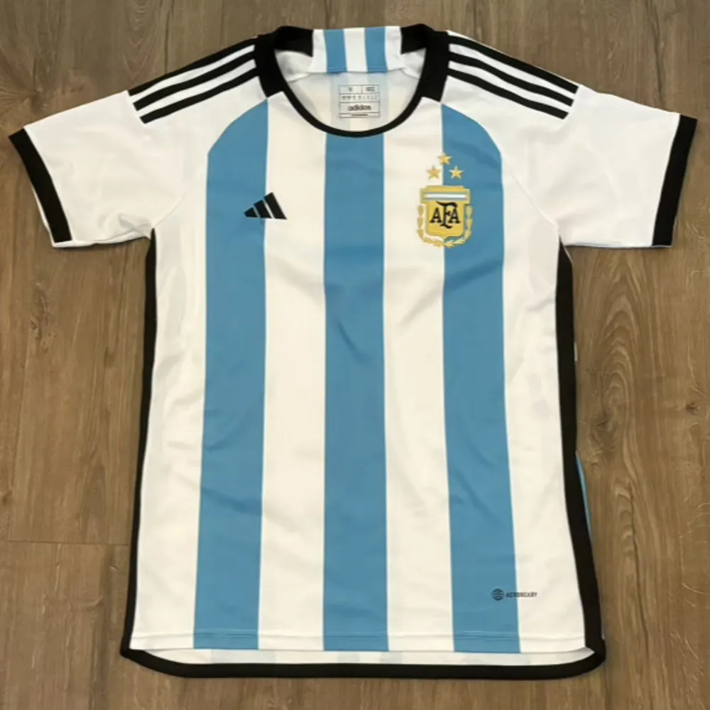 Säljer en snygg Argentina fotbollströja från Adidas i storlek S. Tröjan har de klassiska blå och vita ränderna med AFA-emblemet på bröstet. Den är kortärmad och har svarta detaljer vid krage och ärmslut. Perfekt för fotbollsfans!. T-shirts.