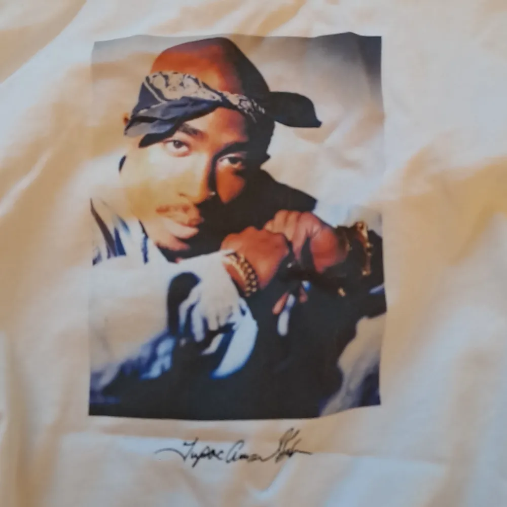 Säljer en vit T-shirt från H&M med ett coolt tryck av 2Pac på framsidan. T-shirten har en loose fit och är perfekt för en avslappnad stil. Materialet känns mjukt och skönt mot huden. Perfekt för alla 2Pac-fans där ute!. T-shirts.