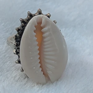 Ring med snäckskal och silverdetaljer - Unik ring med ett stort snäckskal i centrum, omgiven av detaljerade silverfärgade kanter som ger en rustik känsla. Perfekt för att ge en bohemisk touch till din outfit. Snäckskalet har en naturlig vit färg med en mjuk orange ton inut.