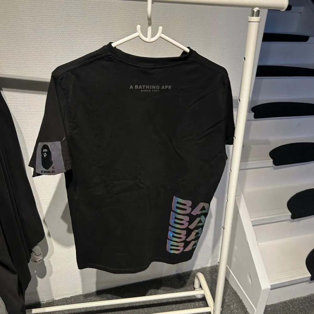 Säljer en svart t-shirt från A Bathing Ape med deras ikoniska logga i ett coolt, färgglatt mönster på framsidan. T-shirten har korta ärmar och texten 'BY BATHING APE' under loggan. På sidan finns det flerfärgad text 'APE' som ger en extra touch. Perfekt för streetwear-stilen!                                   Är storlek L men upplevs mindre. T-shirts.