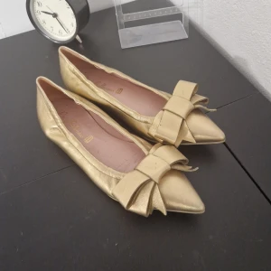 Guldiga ballerinaskor med rosett - Säljer ett par eleganta guldiga ballerinaskor med en stor rosett på tån. Perfekta för festliga tillfällen eller när du vill känna dig extra glamorös. Skorna har en spetsig tå och är i syntetmaterial.