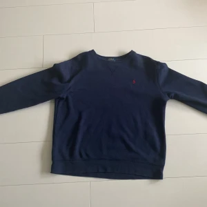 Mörkblå tröja från Polo Ralph Lauren - Säljer en snygg mörkblå tröja från Polo Ralph Lauren. Den har en klassisk design med det ikoniska röda logotypbroderiet på bröstet. Tröjan är långärmad och perfekt för både höst och vinter. Den är i bra skick och redo för en ny ägare!