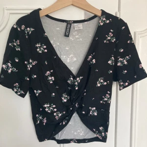 Blommig topp från H&M Divided - Säljer en superfin blommig topp från H&M Divided. Den är svart med små rosa och vita blommor över hela tyget. Toppen har en v-ringad design och en knutdetalj framtill som ger en snygg twist. Den är kortärmad och perfekt för både vardag och fest! Står storlek XS men passar fint till S också💕