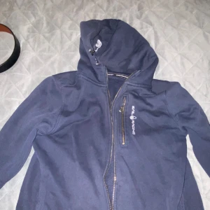 Mörkblå jacka från Sail Racing - Säljer en mörkblå jacka från Sail Racing. Den har en dragkedja framtill och en huva med logga. Perfekt för kyliga dagar och har en sportig look.