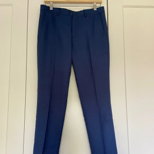 Blå kostymbyxor från Jack & Jones - Säljer ett par stilrena blå kostymbyxor från Jack & Jones i super slim passform. Byxorna har en klassisk design med dragkedja och knappar framtill samt fickor både fram och bak. Perfekta för både formella tillfällen och kontoret.