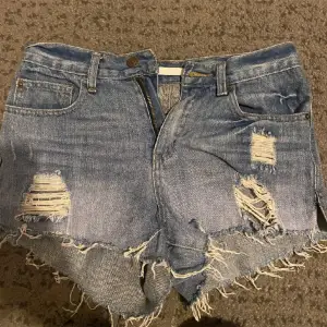 Säljer ett par coola blå jeansshorts med slitningar och fransiga kanter. De har en klassisk femficksdesign och knappar i midjan. Perfekta för en avslappnad sommarlook!