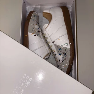 Vita sneakers med färgstänk Från Maison Margiela - Säljer ett par vita sneakers från Maison Margiela i nytt skick. De har en unik design med färgstänk i blått, rött och svart över tåpartiet. Skorna har en klassisk siluett med snörning och en brun sula. Perfekta för att ge din outfit en konstnärlig touch! PERFEKT JULKLAPP FÖR TÅNÅRINGAR 