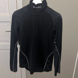 Svart långärmad tröja från Bauer med halsskydd - Säljer en svart långärmad underställströja från Bauer i bra skick. Perfekt för hockey träning. Tröjan har halsskydd och snygga detaljer i grått längs sidorna. Materialet är mjukt och stretchigt för bästa komfort. 
