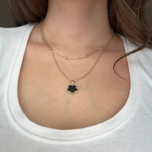 Guldhalsband med svart blomma - Snyggt guldhalsband med en svart blomformad berlock. Halsbandet har en tunn kedja med små guldkulor som detaljer. Perfekt för att ge en elegant touch till vilken outfit som helst. Passar både till vardags och fest!