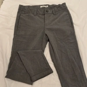Grå byxor från Name It - Säljer ett par stilrena ljusgrå byxor från Name It i storlek 152. Super mjuka och sköna chinos. De har en klassisk design med knapp och dragkedja framtill samt hällor för bälte. På insidan finns det elastisk dragsko för att justera midjan.  Perfekta för både vardag och festliga tillfällen. Byxorna är i mycket bra skick. 