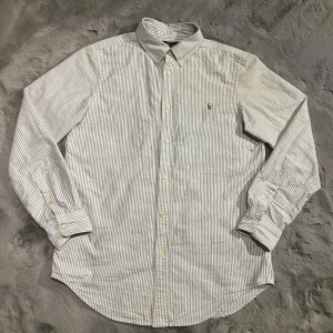 Randig skjorta från Ralph Lauren - Säljer en klassisk vit och blå randig skjorta från Ralph Lauren i storlek S. Skjortan är i bomull och har långa ärmar med knappar framtill. Perfekt för både vardag och festliga tillfällen. Den har en broderad logga på bröstet som ger en stilren touch. Passar bra till både jeans och kostymbyxor!