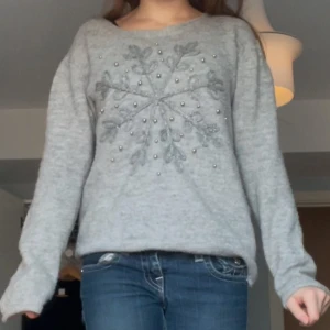 Grå tröja med snöflingemönster - Mysig grå tröja med ett stort snöflingemönster på framsidan. Perfekt för kyliga dagar med sin bekväma passform och långa ärmar. Passar bra till jeans för en avslappnad look.