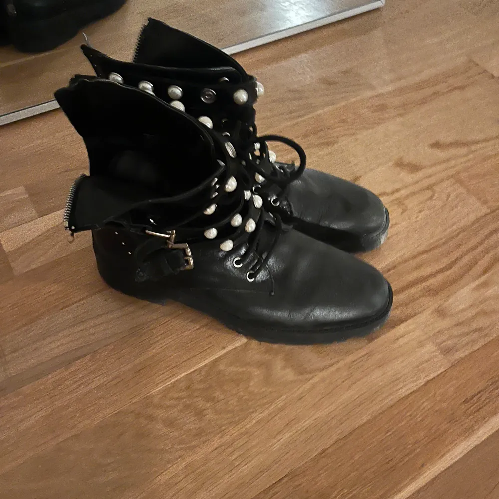 Säljer ett par svarta boots i skinn med coola pärldetaljer och snörning. De har även en dragkedja på sidan och ett bälte med spänne för extra stil. Perfekta för höst och vinter, och ger en edgy look till vilken outfit som helst. Använda men i bra skick.. Skor.