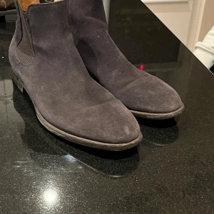 Marinblå mocka chelsea boots - Säljer ett par stilrena marinblå chelsea boots i mocka. De har en klassisk design med elastiska paneler på sidorna för enkel på- och avtagning. Skorna är i bra skick med lite slitage på sulorna, perfekta för både vardag och fest. 