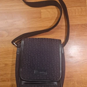 Svart axelväska/becknarväska från Calvin Klein - Snygg svart axelväska från Calvin Klein med justerbar rem. Väskan har ett diskret CK-mönster och en praktisk dragkedja inuti för säker förvaring. Två stycken innerfickor. Perfekt för vardagsbruk eller en kväll ute. Den är i mycket bra skick och redo att användas direkt!