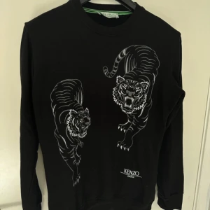 Svart tröja med tigertryck från Kenzo - Säljer en svart långärmad tröja från Kenzo med coolt tigertryck på framsidan. Tröjan är i bra skick och perfekt för både höst och vinter. Den har en normal passform och är gjord av mjukt material som känns skönt mot huden. Perfekt för dig som vill ha en stilren men ändå iögonfallande look!